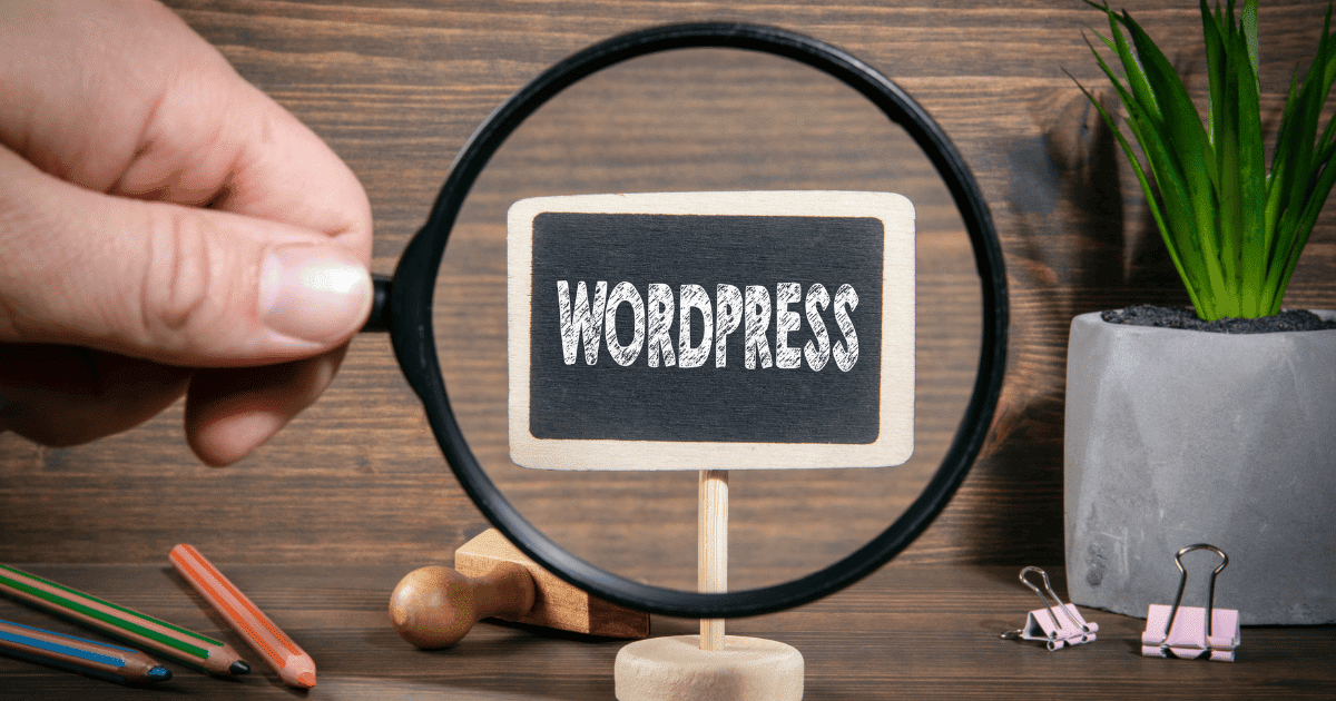 Una mano sosteniendo una lupa que enfoca un pequeño cartel con la palabra WordPress, rodeado de material de oficina, representando la investigación sobre cómo saber los plugin que usa una web.