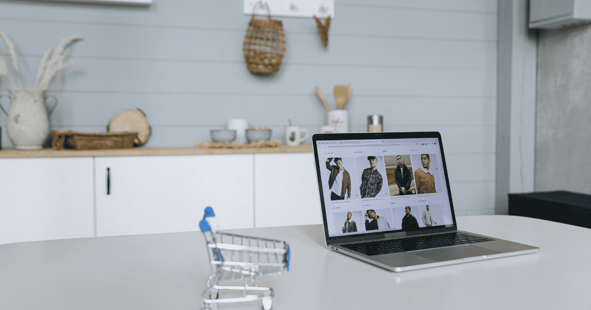 Un ordenador portátil abierto mostrando una tienda online de ropa en una mesa blanca junto a un pequeño carrito de la compra metálico.